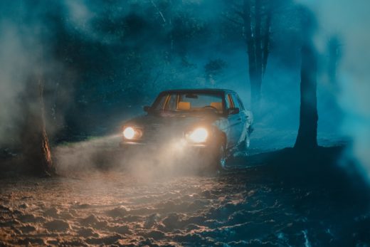 A car in a misty forest at night with visible headlight beams.