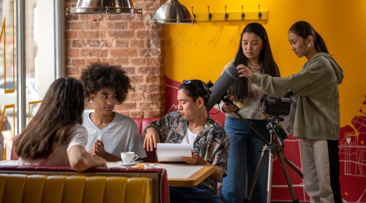 A small film shoot with a young cast and crew works in a brightly-coloured restaurant interior.