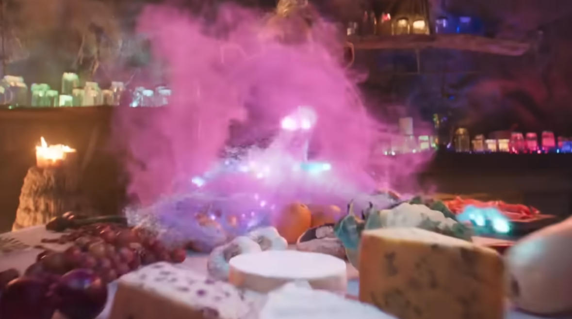 The BFG character looks over a table as it is filled with food by a purplish cloud of magical dust.